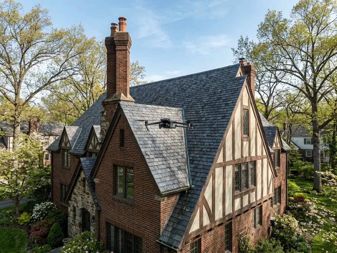 Small quadcopter drone hovering above a steep slate-gray shingle roof with a brick chimney on a Tudor house